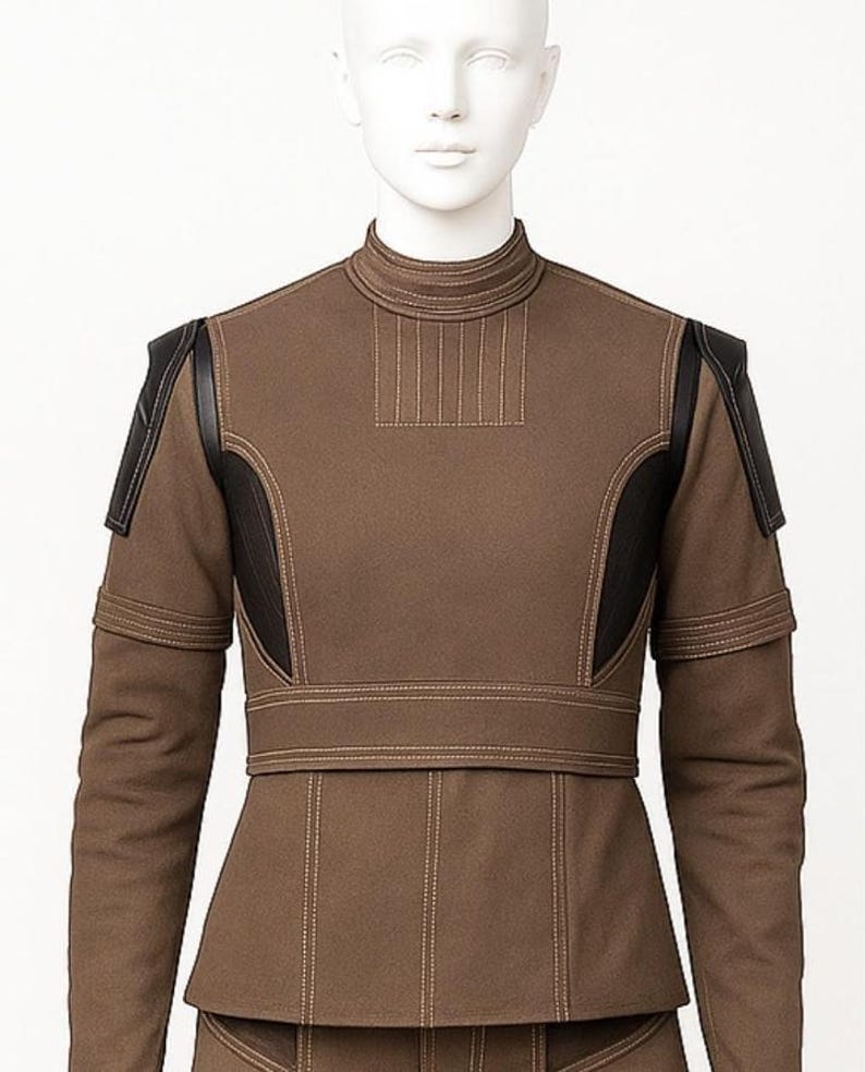 May include: A brown jacket with black leather panels on the shoulders and sides. It features a high collar, vertical stitching, and a fitted waist. The sleeves have a double-layered design. The jacket is displayed on a mannequin.