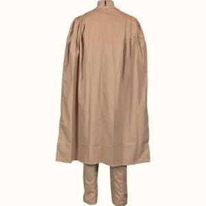 May include: A tan-colored, two-piece outfit featuring a long, flowing cape-like top and matching pants. The top has a high collar and a cape that drapes down the back. The pants are straight-legged. The outfit is made of a smooth, solid-colored fabric.