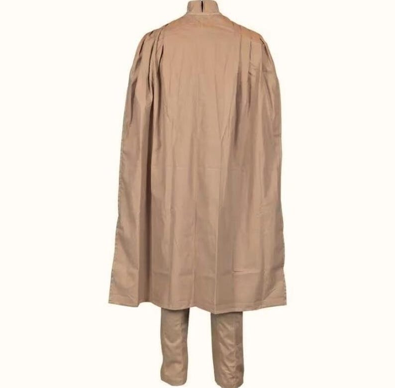 May include: A tan-colored, two-piece outfit featuring a long, flowing cape-like top and matching pants. The top has a high collar and a cape that drapes down the back. The pants are straight-legged. The outfit is made of a smooth, solid-colored fabric.