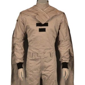 May include: Tan-colored costume with a cape, featuring a high collar and black accents. The outfit includes a long-sleeved top with two black rectangular patches and matching pants. Black cuffs are visible at the wrists.