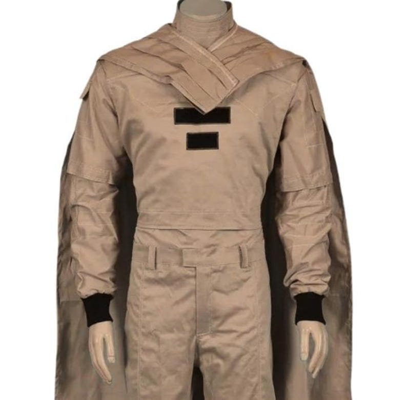 May include: Tan-colored costume with a cape, featuring a high collar and black accents. The outfit includes a long-sleeved top with two black rectangular patches and matching pants. Black cuffs are visible at the wrists.