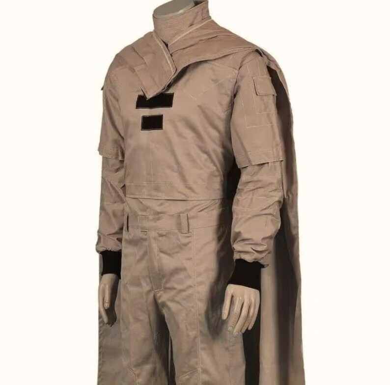 May include: A tan-colored costume with a high collar and cape. The outfit features black accents on the chest and black cuffs. The costume includes a long-sleeved jacket and matching pants. The mannequin is wearing black gloves.
