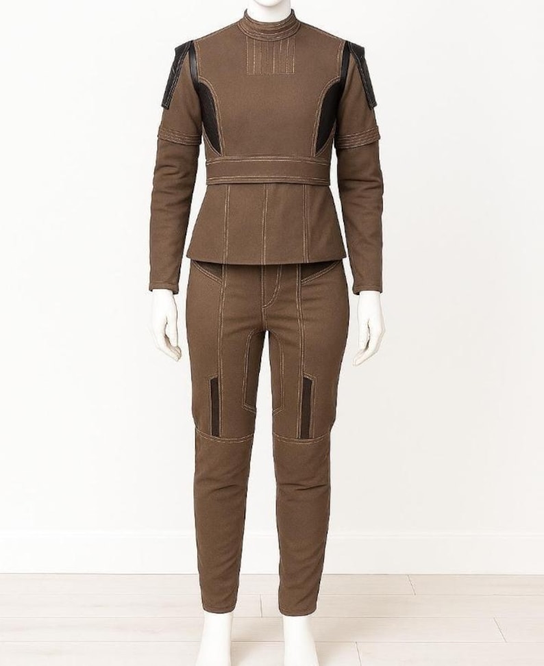 May include: A brown two-piece outfit displayed on a mannequin. The long-sleeved jacket features black shoulder accents and a high collar. The fitted trousers and jacket have visible stitching. The outfit is a solid brown colour.