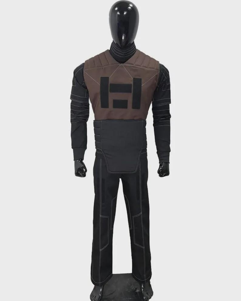 K&ouml;nnte beinhalten: Schwarzes und braunes taktisches Outfit auf einer Schaufensterpuppe. Das Outfit umfasst eine braune Weste mit schwarzem geometrischen Design, ein schwarzes Langarmshirt, schwarze Hosen mit wei&szlig;en N&auml;hten und schwarze Handschuhe. Die Schaufensterpuppe hat einen schwarzen Kopf.