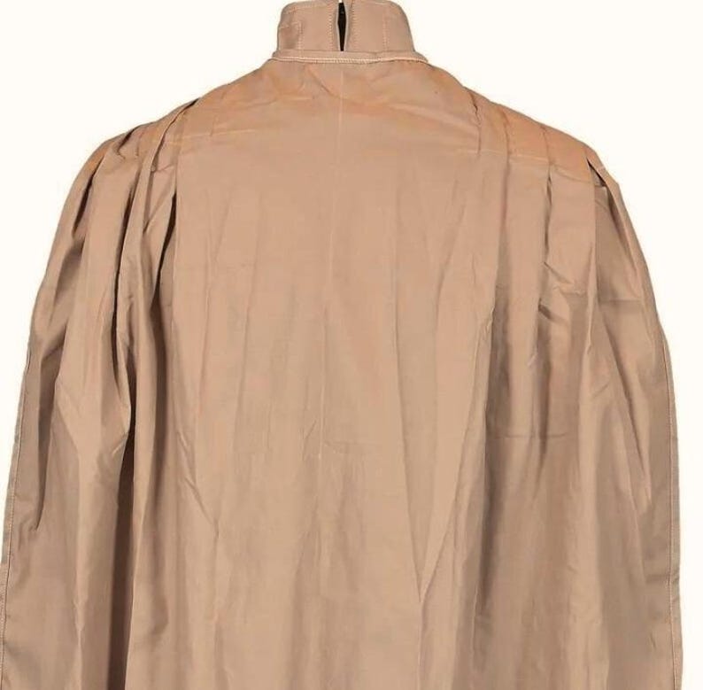 May include: A tan-colored cape with a high collar and a pleated design on the shoulders. The cape is made of a lightweight fabric and has a flowing silhouette. The back view of the cape is shown against a white background.
