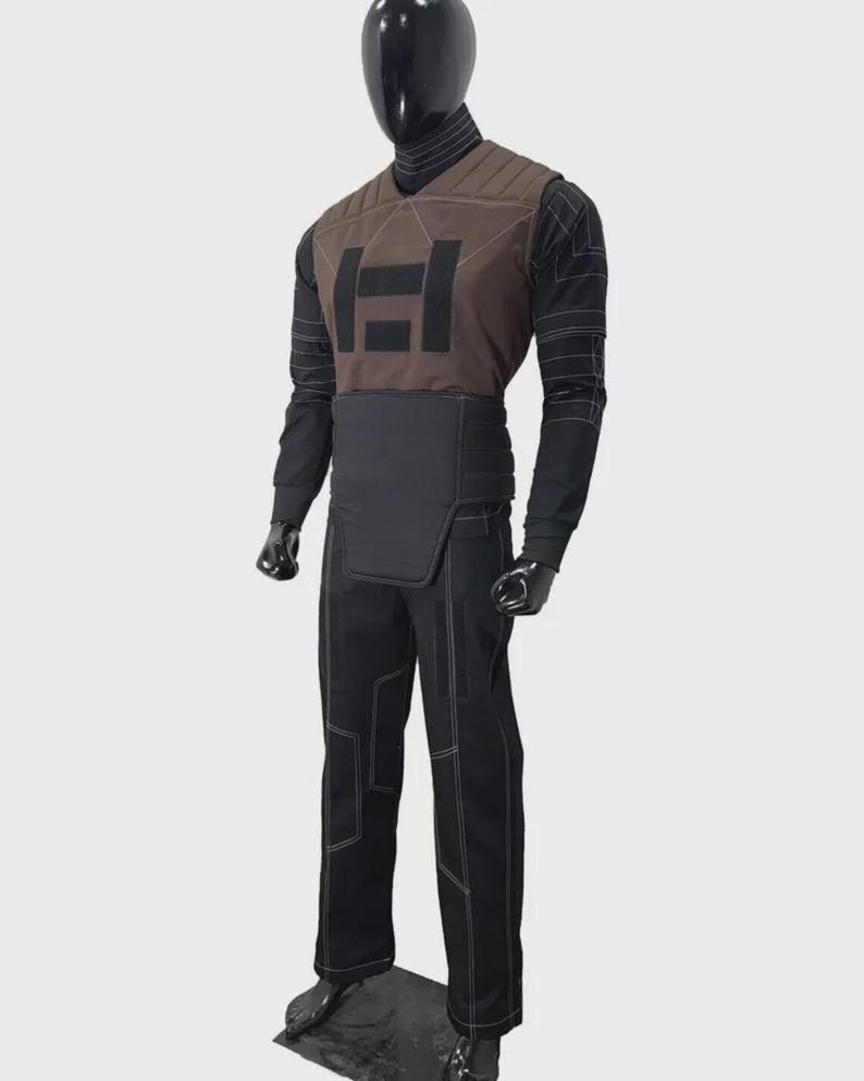 K&ouml;nnte beinhalten: Ein Ganzk&ouml;rperkost&uuml;m, das auf einer Schaufensterpuppe pr&auml;sentiert wird. Das Kost&uuml;m umfasst eine braune Weste mit schwarzem Logo, ein schwarzes Langarmshirt, eine schwarze Hose mit wei&szlig;en N&auml;hten und einen schwarzen G&uuml;rtel. Die Schaufensterpuppe ist schwarz.