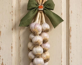 May include: A decorative hanging bundle of dried garlic bulbs tied together with twine and topped with a large olive green fabric bow. The center of the bow features a small rectangular patch with an embroidered orange pumpkin design.