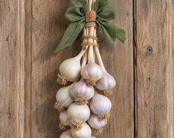 May include: A decorative hanging bundle of artificial garlic bulbs with off-white and light purple skins. The stems are tied together with natural twine and topped with a green fabric bow featuring a small orange maple leaf accent.