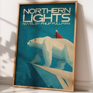Puede incluir: Póster enmarcado de la portada del libro "Luces del Norte" de Philip Pullman. Muestra un oso polar en un acantilado, con una figura roja sobre su espalda. El fondo es azul y el título es dorado. El póster está en un marco de madera.