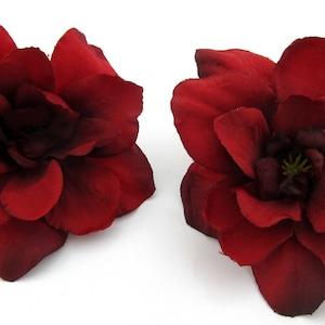May include: Two deep red artificial flower hair clips. Each clip features a large, multi-petaled bloom with a dark center. The flowers are set against a white background, highlighting their rich color and texture. These are ideal for adding a touch of elegance to any hairstyle.