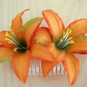 May include: A pair of orange lily flowers with green centers are attached to a white plastic comb. The flowers are made of fabric and have a realistic appearance.