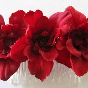 May include: A floral hair comb featuring three large, deep red magnolia blossoms. The flowers have a velvety texture and are attached to a clear plastic comb. The comb is designed for securing hair in place.