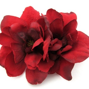 May include: A deep red, artificial flower with multiple layered petals. The flower has a dark center and is set against a white background. The petals have a textured appearance, suggesting a fabric material.