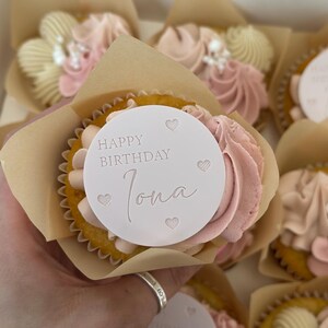 May include: Cupcakes with pink buttercream frosting and a white circular topper that reads "Happy Birthday Iona" with heart accents. The cupcakes are in brown paper wrappers and arranged in a box. The frosting is a light pink colour.
