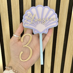 May include: A layered cake topper featuring a light purple seashell shape with the name Blousiee in gold script lettering. A separate gold number 3 is attached to a light blue vertical stick, creating a decorative piece for a celebration.
