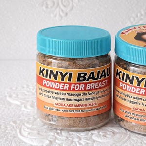 以下が含まれることがあります： 透明なプラスチックジャー、ターコイズブルーの蓋付き、茶色の粉末入り。ラベルには太字で「KINYI BAJAU POWDER FOR BREAST」と書かれています。追加のテキストが下に印刷されています。