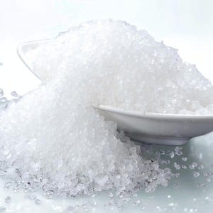 Peut inclure: Gros plan sur un tas de cristaux blancs grossiers, possiblement du sel ou du sucre, débordant d'une coupelle blanche. Les cristaux ont une qualité translucide, captant la lumière. L'arrière-plan est blanc et flou.