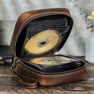 Puede incluir: Un estuche de CD de cuero marrón, abierto para revelar varios CD en fundas transparentes. El estuche tiene cierre de cremallera y una correa de muñeca. Los CD son dorados y el estuche está sobre una superficie de madera.