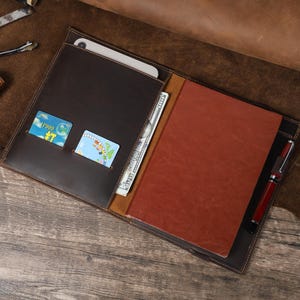 May include: Open brown leather portfolio with a red notebook, pen, and pockets for cards and cash. The portfolio is open, revealing a tablet and a stack of US dollar bills.