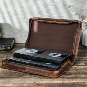 May include: A brown leather CD case, open to reveal multiple CD sleeves. The case has a zipper closure and a stitched design. The CDs are visible inside the sleeves. The case is on a wooden surface.