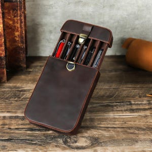 Puede incluir: Un estuche de cuero marrón oscuro, abierto para revelar varios bolígrafos. El estuche tiene forma rectangular con una tapa con bisagras y un cierre dorado. El interior está forrado con una tela marrón suave, y los bolígrafos se guardan en ranuras individuales.
