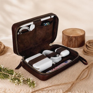 May include: Open brown leather travel case with a zip, containing a white charger, earbuds, and a stylus. The case has multiple compartments and a brown corduroy lining. A small wooden log and rope are in the background.