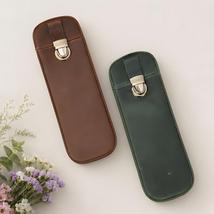 Puede incluir: Dos estuches de cuero para bolígrafos, uno marrón y otro verde oscuro. Cada estuche tiene forma rectangular con la parte superior redondeada y un cierre de metal. Los estuches están sobre una superficie blanca, con un pequeño arreglo floral.