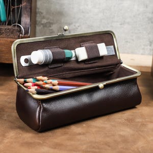 May include: A brown leather pencil case with a metal clasp, open to reveal coloured pencils, a tube of paint, an eraser, and a ruler. The case has a dark brown interior and a vintage aesthetic.
