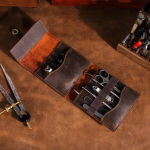 May include: A brown leather manicure set, open and laid flat. The set includes various grooming tools such as scissors, nail clippers, and cuticle pushers. The interior is lined with a lighter brown material, and the set is secured with a leather strap.
