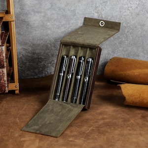 Puede incluir: Un estuche para bolígrafos de cuero marrón oscuro con forro interior de ante verde oliva, diseñado para cuatro unidades. El estuche cuenta con una solapa con un botón de presión de metal plateado, albergando cuatro bolígrafos negros.