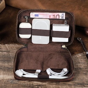 May include: A brown leather zippered organizer case featuring an interior with elastic bands holding white electronic accessories, including a charging block, a wireless charger, and earbuds, with white cables and a stylus stored in bottom pockets.