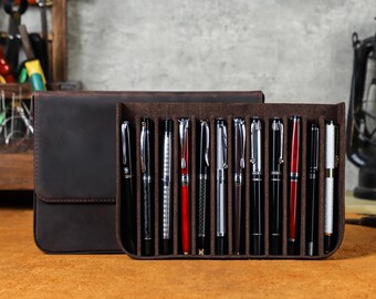 May include: A brown leather pen case with a flap closure, next to a pen holder containing a collection of fountain pens. The pens are black, silver, and red, with various designs. The case and holder sit on a wooden surface.