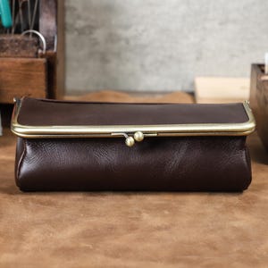 Puede incluir: Un bolso de mano de cuero marr&oacute;n oscuro con cierre de broche de lat&oacute;n. El bolso rectangular tiene una forma redondeada y una est&eacute;tica vintage. El cierre presenta dos peque&ntilde;os detalles redondos de lat&oacute;n. El bolso de mano se muestra sobre una superficie marr&oacute;n.
