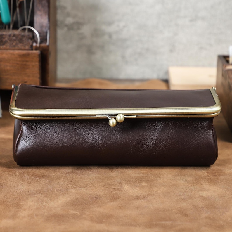 Puede incluir: Un bolso de mano de cuero marr&oacute;n oscuro con cierre de broche de lat&oacute;n. El bolso rectangular tiene una forma redondeada y una est&eacute;tica vintage. El cierre presenta dos peque&ntilde;os detalles redondos de lat&oacute;n. El bolso de mano se muestra sobre una superficie marr&oacute;n.