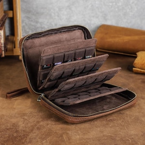 May include: A brown leather zipped storage case featuring multiple interior tiered panels with individual slots designed for organising small items. The case has a wrist strap attached to the side and a soft, dark brown suede-like interior lining.
