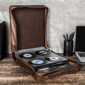 May include: A brown leather vinyl record carrying case, open to reveal several vinyl records in protective sleeves. The case has a zipper closure and is sitting on a wooden surface. A black pencil holder and laptop are in the background.