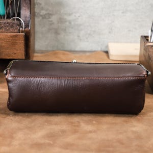 Puede incluir: Un estuche de l&aacute;pices de cuero marr&oacute;n oscuro con un cierre de metal. El estuche rectangular tiene costuras visibles en hilo marr&oacute;n claro. El estuche est&aacute; sobre una superficie marr&oacute;n.