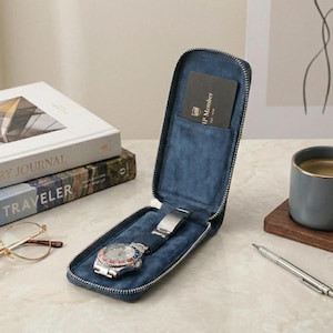 Puede incluir: Un estuche de viaje azul para reloj con cremallera plateada, abierto para revelar un reloj de pulsera con una correa azul y un cierre plateado. Una tarjeta negra está en el bolsillo. El estuche está sobre una superficie de mármol.