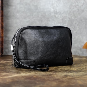 May include: A black leather clutch with a silver zipper closure and a wrist strap. The rectangular bag has rounded corners and a smooth texture. The zipper runs along the top and around the sides of the bag.
