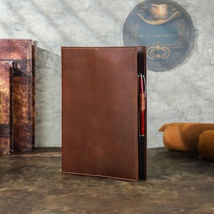 May include: A brown leather portfolio with a pen holder and a red pen. The portfolio is standing upright on a wooden surface. The background includes a decorative wall hanging and a stack of books.