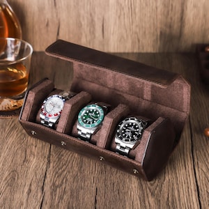 May include: A brown leather watch case with three watches. The watches have silver bands and different colored faces: red, white, and blue; green; and black. The case is open, revealing a soft, brown interior.