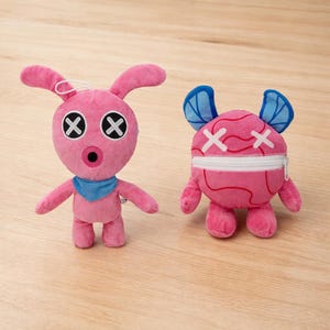 Puede incluir: Dos peluches rosas. Uno tiene orejas de conejo, ojos tachados y una bufanda azul. El otro tiene elementos azules en forma de ala, ojos tachados y una cremallera. Ambos están sobre una superficie de madera clara.