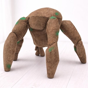 May include: A brown, plush, six-legged creature with green accents. The handmade toy has a textured appearance and is posed on a white surface. The creature's body is a rounded shape with articulated legs.