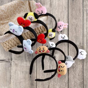 May include: A collection of black headbands adorned with plush cartoon characters in various colors. The characters include a pink bunny, yellow dog, red heart, blue koala, and other cute designs. The headbands are displayed on a wooden surface.