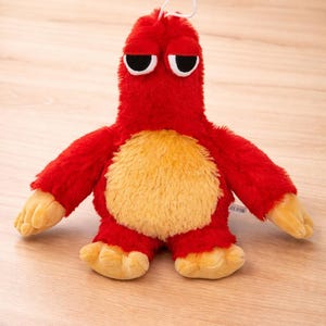 Puede incluir: Un peluche rojo con forma de criatura con vientre, brazos y pies amarillos. Tiene ojos negros y un lazo blanco. El juguete está sobre una superficie de madera de color marrón claro.