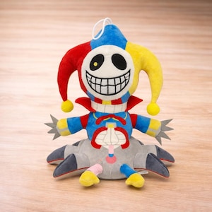 Puede incluir: Muñeco de bufón de peluche con una cara sonriente, que lleva un sombrero rojo, azul y amarillo. El muñeco tiene un cuello rojo, un cuerpo azul y una parte inferior del cuerpo gris con múltiples apéndices. El muñeco tiene un lazo de cuerda blanco en la parte superior.