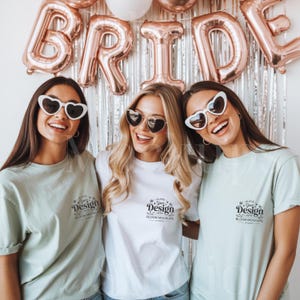 May include: Three smiling people wearing heart-shaped sunglasses and t-shirts. The shirts have a small logo on the chest. Behind them are rose gold foil balloons spelling out "BRIDE" and a silver tinsel backdrop.