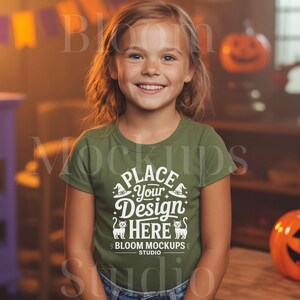 Op de afbeelding: Een jong meisje draagt een olijfgroen t-shirt met de tekst "Place Your Design Here" in het wit. Het ontwerp bevat heksenhoeden en katten silhouetten, met "Bloom Mockups Studio" eronder. De achtergrond heeft Halloween-decoraties.