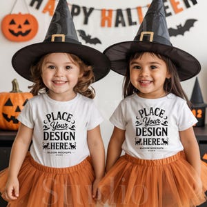 Op de afbeelding: Twee jonge kinderen in Halloween-kostuums. Ze dragen zwarte heksenhoeden met gouden gespen, witte t-shirts met de tekst "PLACE YOUR DESIGN HERE" en oranje tule rokken. Halloween-decoraties op de achtergrond.