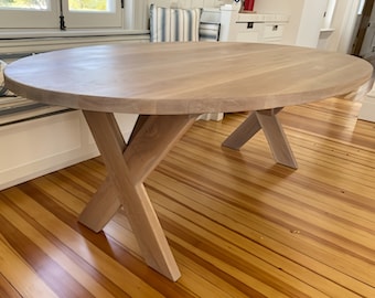 May include: A round wooden table with a light wood finish and a cross-shaped base. The table is sitting on a wood floor.